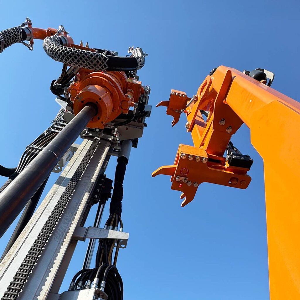 Upward view of orange hydraulic components and drill head on a Fraste ML MAX rig, highlighting precision engineering against a clear blue sky.